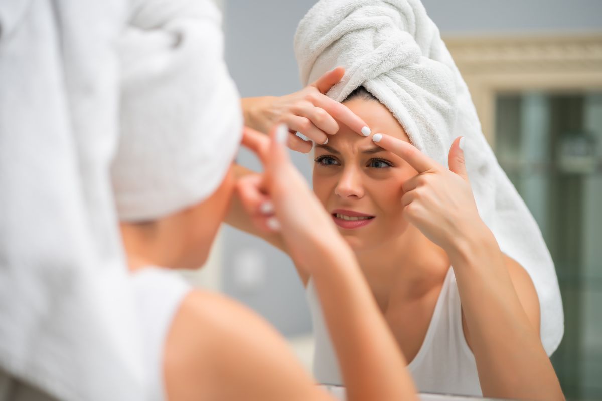 Kobieta stojąca przed lustrem, z ręcznikiem na głowie, walcząca z niedoskonałościami na twarzy.